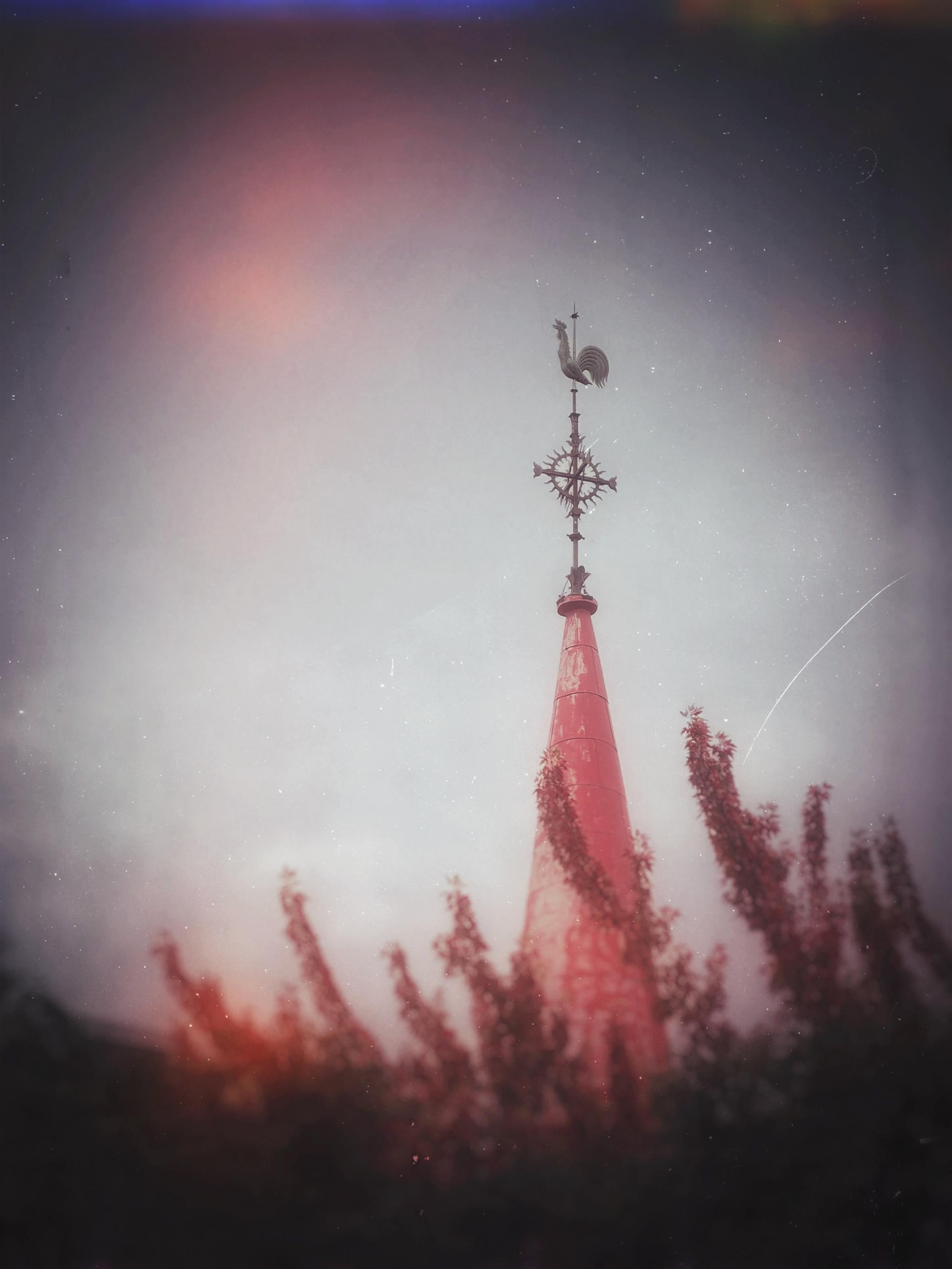 This image features a tall, slender red spire rising above a foreground of blurred, reddish-brown foliage. At the top of the spire, there is an ornate metal cross topped with a rooster weather vane. The background is a soft, muted sky with a vintage, slightly distressed texture, giving the scene a somewhat moody or nostalgic feel.