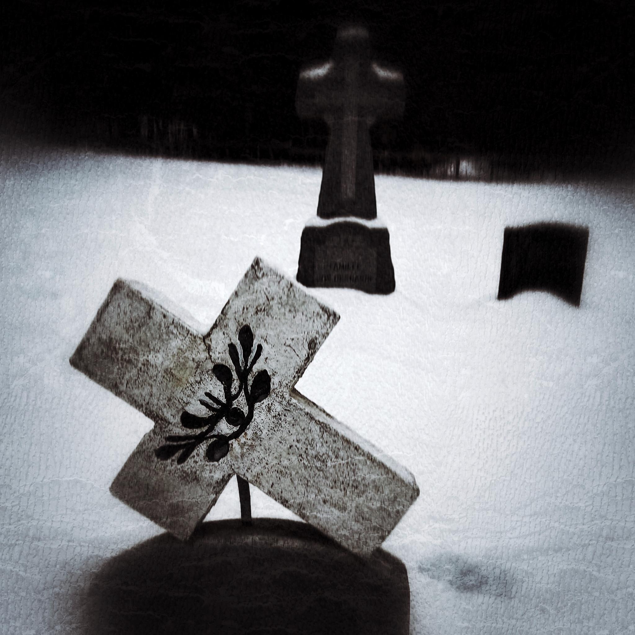 This image shows a snowy graveyard scene. In the foreground, there is a weathered stone cross tilted to the side, featuring a dark, leafy design carved into its center. In the background, partially obscured by the snow and shadows, are two other grave markers, including one upright cross.