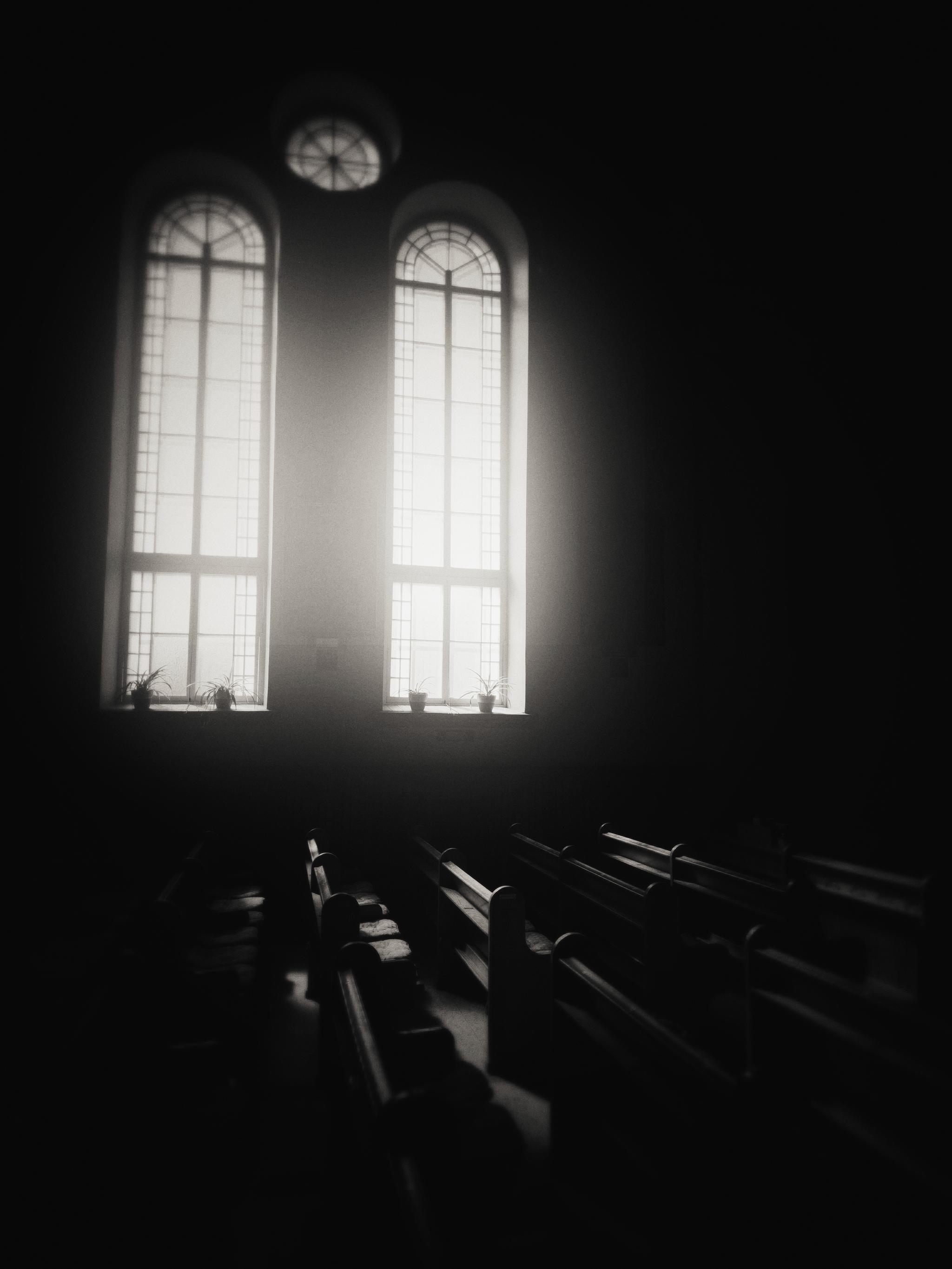 This black-and-white photo captures a quiet, contemplative scene inside what appears to be a church or chapel. Two tall, arched windows with a grid pattern allow bright light to stream into the dim interior, illuminating the tops of the wooden pews below. Small potted plants sit on the windowsills, adding a subtle touch of life to the serene, shadowy space. The high contrast between the bright windows and the dark pews creates a dramatic and peaceful atmosphere, perfect for a caption about stillness, reflection, or quiet moments.
