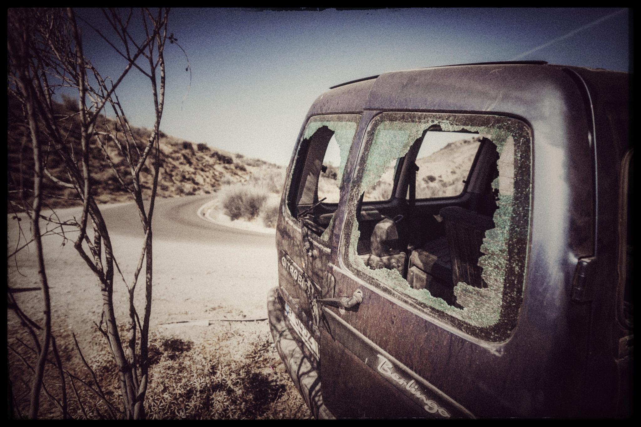 This scene features an abandoned, dark-colored Citroën Berlingo van parked on the side of a paved road in a dry, hilly landscape. The vehicle has extensive damage, most notably large holes in its side and rear windows, with shattered glass still clinging to the frames. A bare, spindly tree stands in the foreground to the left, and the overall setting appears desolate and quiet under a clear sky. The van's weathered appearance and the surrounding arid environment suggest the vehicle has been left there for some time. The road curves gently into the distance, flanked by low, scrubby vegetation on the hillsides.