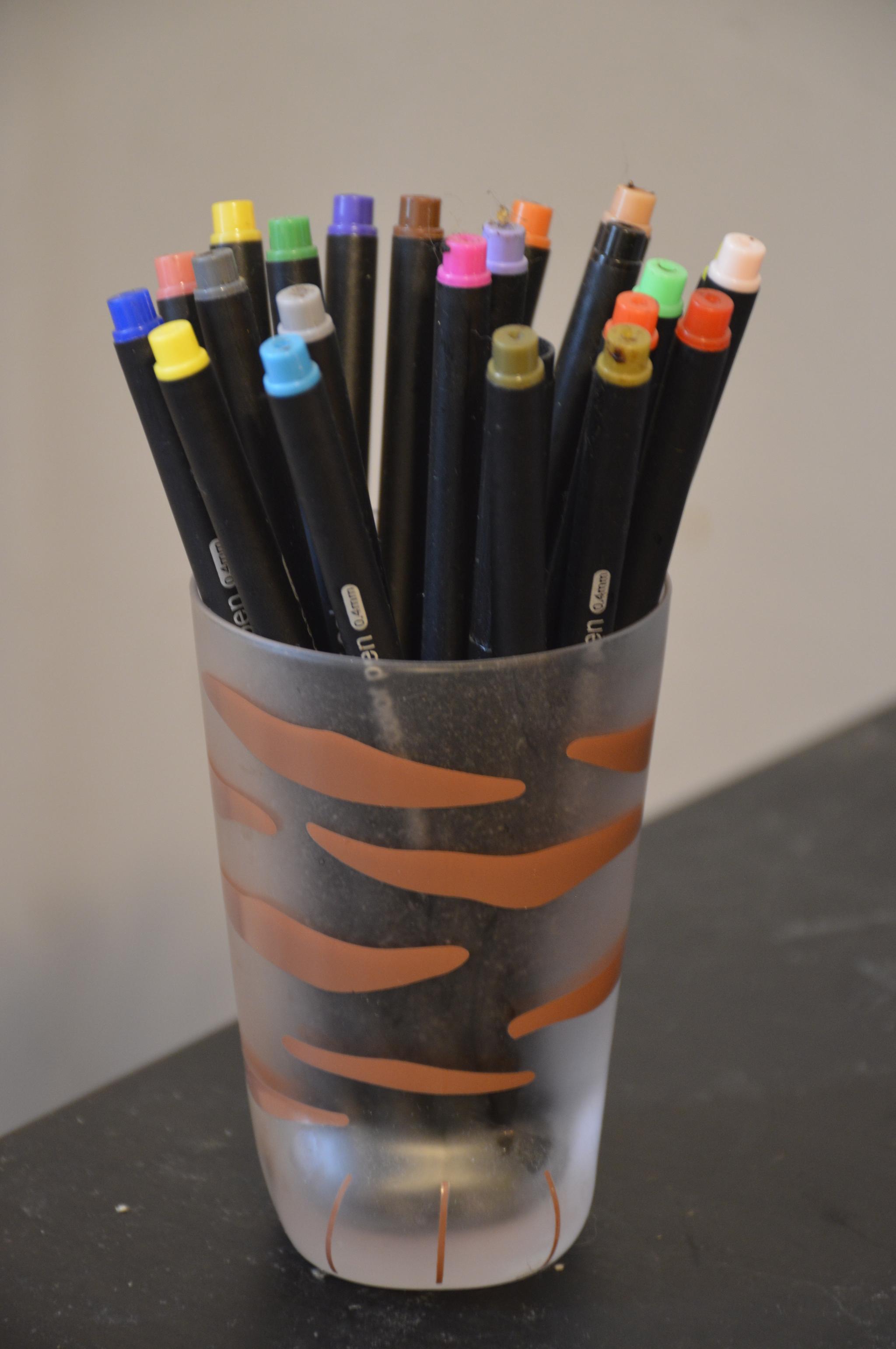 a glass cup shaped like a cat paw. brown broken stripes go up the glass. the glass is holding an assortment of fine tipped colored pens