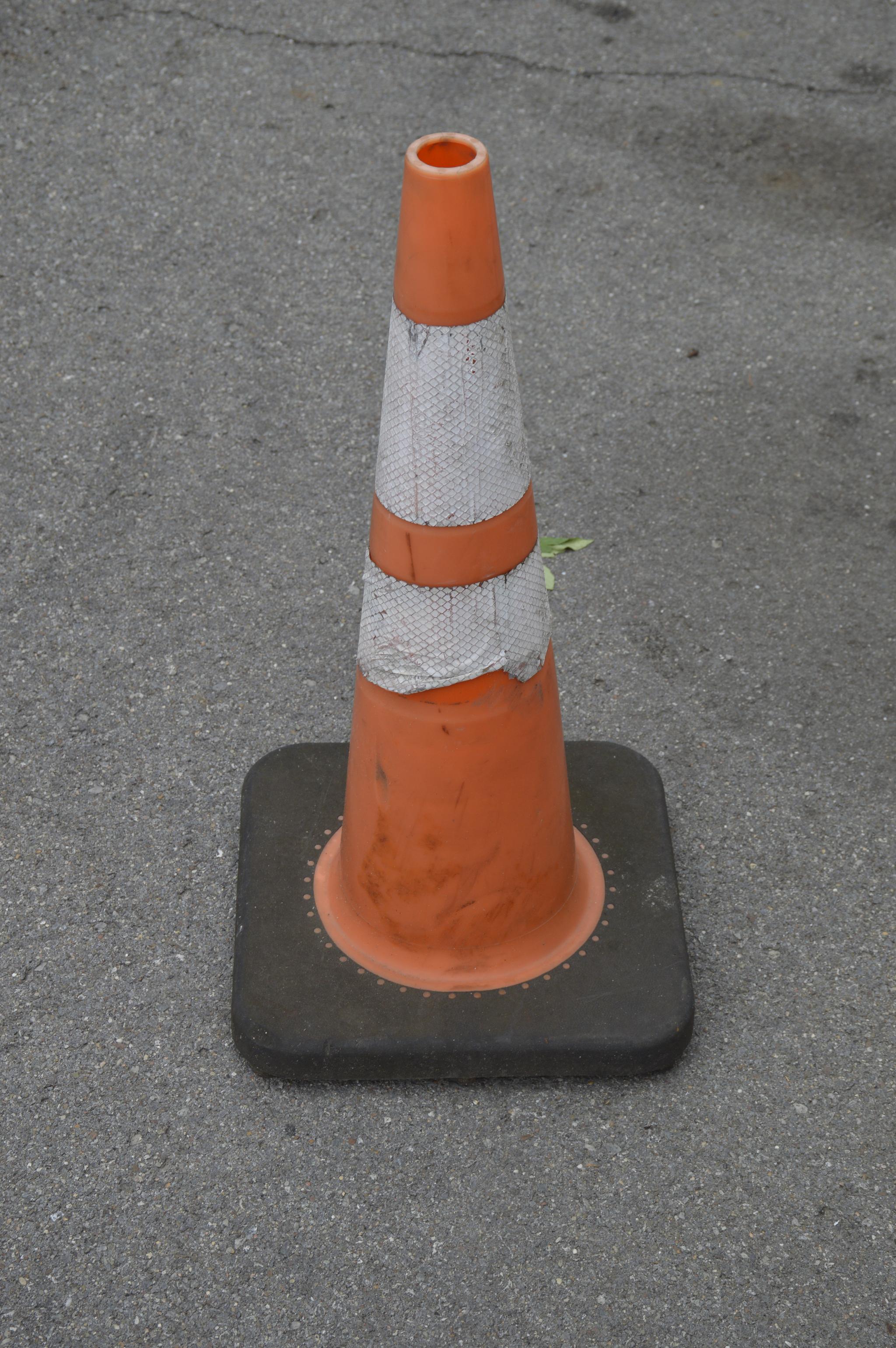 a traffic cone by itself on pavement. a green leaf pokes out from behind it