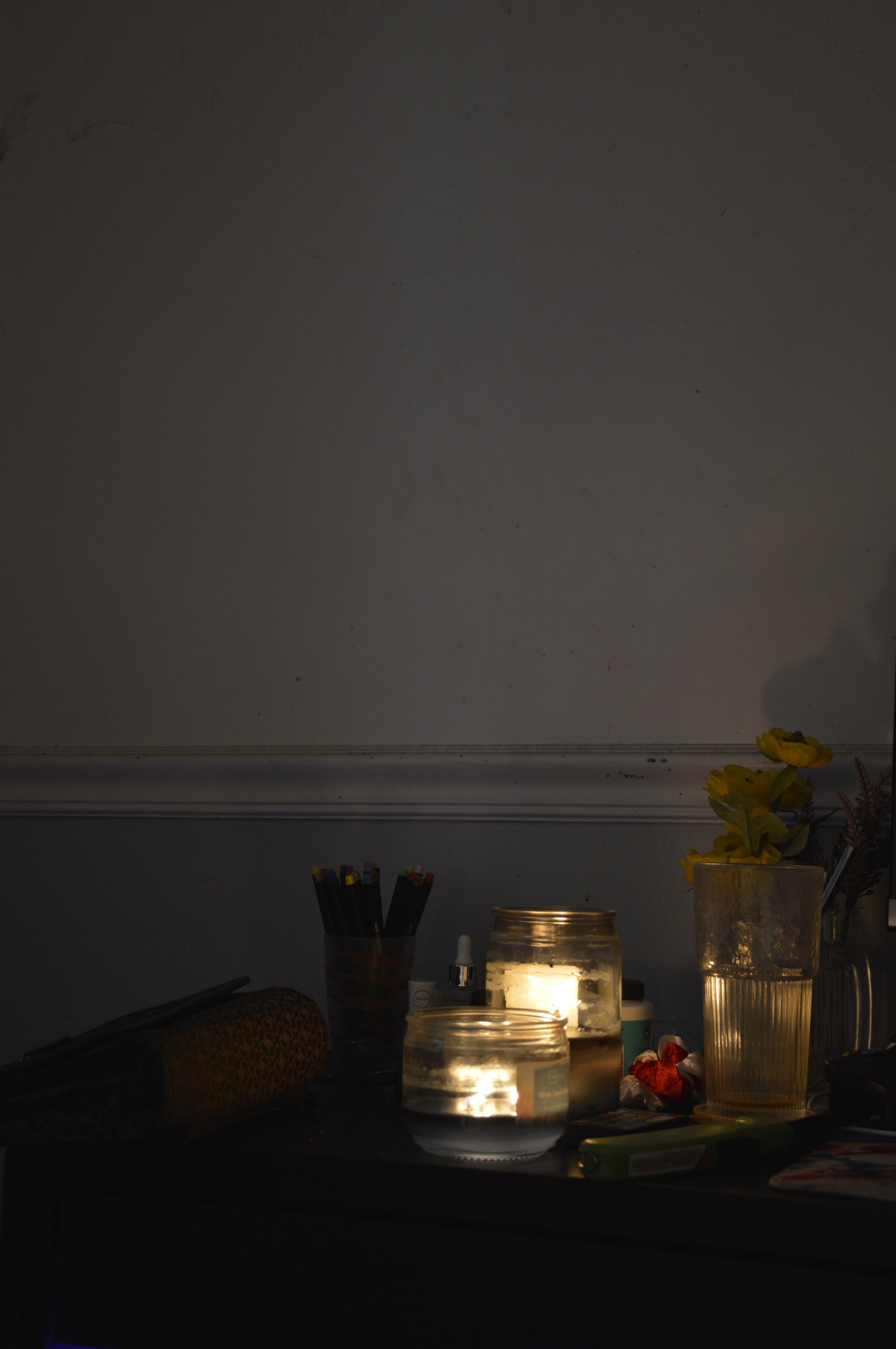 a desk with many items on it including a lit candle, a cup, fake plants, a purse, and colored markers